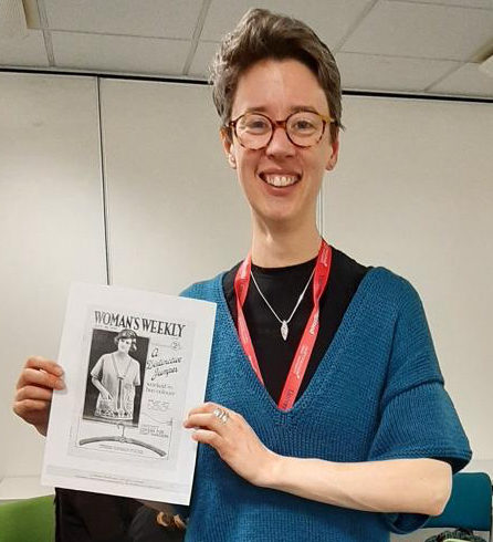 A photograph of Ellie Reed, wearing a blue short-sleeved knitted jumper. She is holding a copy of Woman's Weekly from the 1920s.