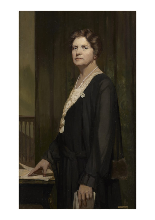 A painting of Lady Rhondda wearing a black dress, a white blouse, and a pearl necklace. She is standing next to a table and holding some papers.