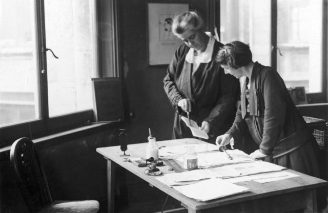 A photo of Helen Archdale (left) and an unnamed person in Time and Tide's offices. They are standing behind a table and looking at some papers. There are stamps and a pot of glue on the table.